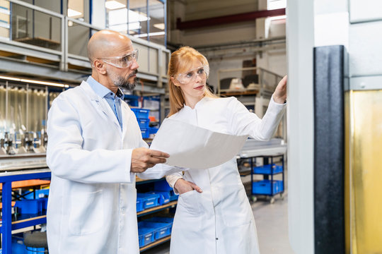 Two technicians wearing lab coats and safety glasses examining machine