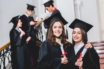selective focus of beautiful girls in graduation caps smiling while holding diplomas near students