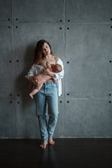 A young mother with a pensive look in white shirt, lowered on her shoulders, and jeans stands at the gray concrete wall holds the baby in her hands and breastfeeds.