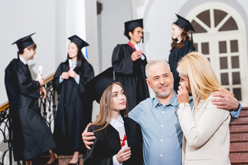 selective focus of attractive girl in graduation cap looking at crying mother while hugging with...