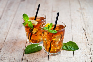Cold iced tea with lemon, mint leaves and ice cubes in two glasses on rustic wooden table.