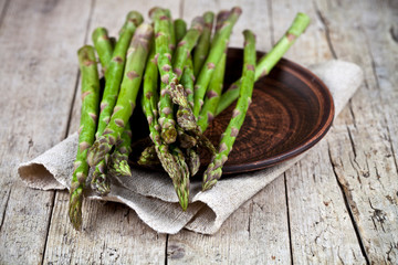 Fresh raw garden asparagus closeup on brown ceramic plate and linen napkin on rustic wooden table background.