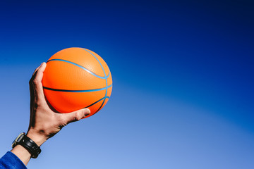 Hand holding an orange basketball ball on blue sky background, invitation to play, copy space free area.