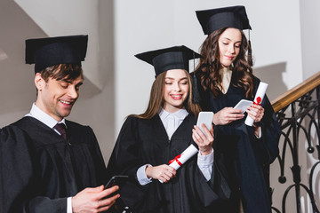 cheerful students in graduation gowns using smartphones while holding diplomas