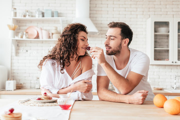 Obraz na płótnie Canvas bearded handsome man feeding girlfriend with berry in kitchen