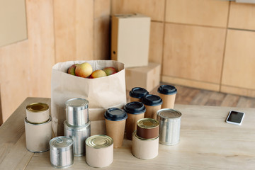 canned food, disposable cups and paper bag with apples on wooden table in charity center