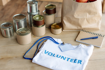 white t-shirt with volunteer inscription, badge,  tins, paper bag and notebook on wooden table