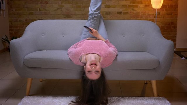 Closeup shoot of young pretty brunette caucasian female watching TV with excitement and lying upside down on the couch in a cozy apartment indoors