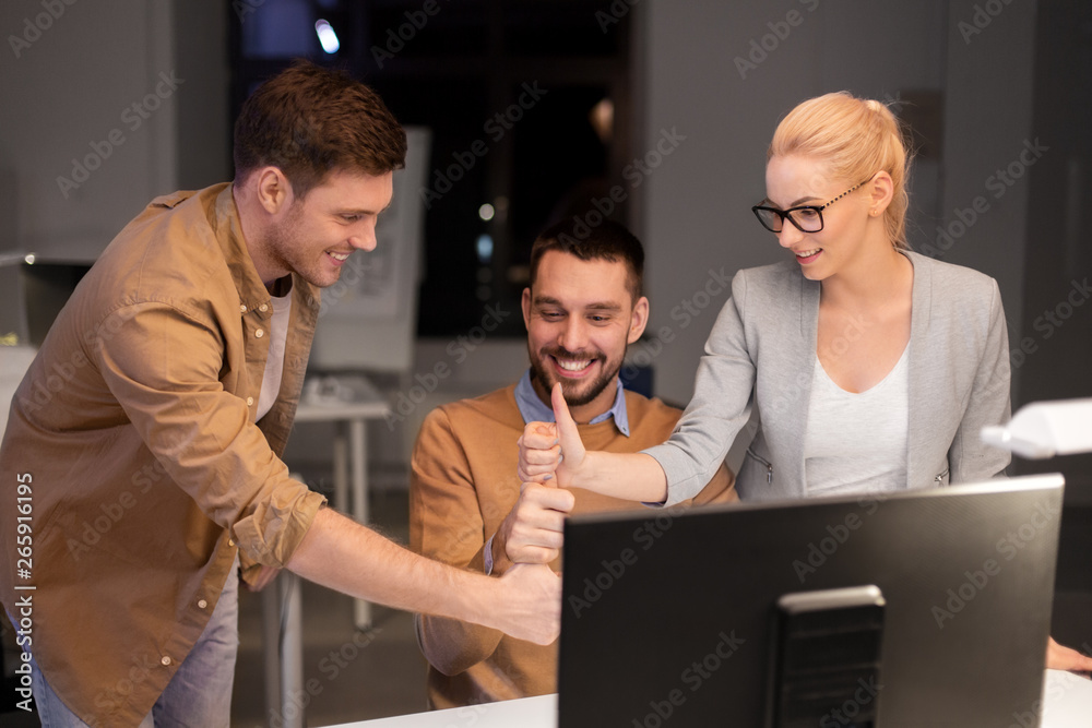 Wall mural business, success and teamwork concept - happy coworkers with computer working late at night office making thumbs up gesture