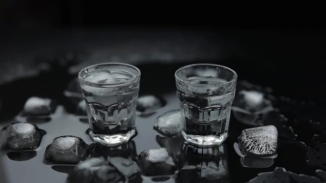 Add ice cubes to shots of vodka in glass against black background. Alcohol drink
