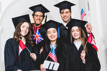 selective focus of cheerful students in graduation gowns holding flags of different countries and taking selfie on smartphone