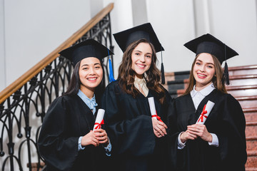 cheerful and beautiful students in graduation caps smiling while holding diplomas in university
