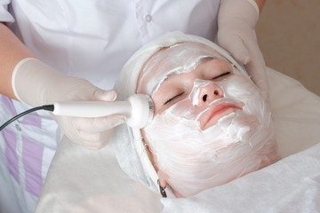 Fototapeta premium The beautician in white gloves holds the electrode of the massage apparatus and smoothes mimic wrinkles on the client's face. Anti-aging effect. Fat woman on the procedure in the cosmetology center.