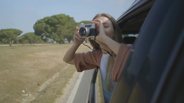 Girl photographing with camera through open car window. Beautiful happy young woman sitting in car and using photo camera through open car window. Photography concept