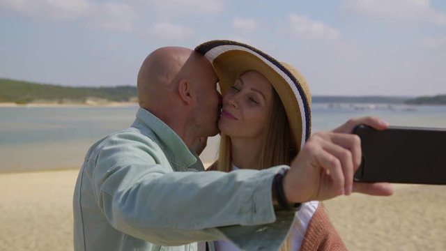 Couple taking selfie with smartphone on beach. Beautiful happy young couple taking pictures with mobile phone and kissing outdoor at sunny day. Technology concept