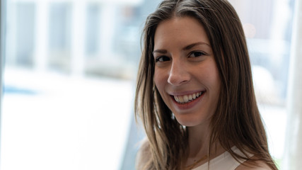 young beautiful long hair brunette woman smiles flirtatiously  by the window light.  She laughs from being in good health. she has a bright white teeth and is very happy. wearing athletic clothing