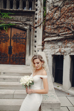 Beautiful bride in a long white dress. Woman with bouquet of flowers. Lady standing near old building