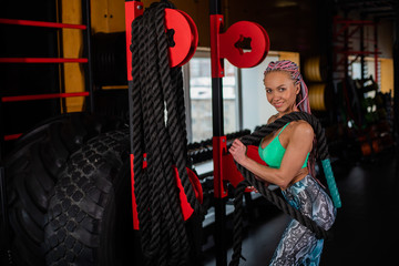 Fototapeta na wymiar Beautiful young woman with colored braids on her head posing in the gym with Battle Rope. Athlete Woman Doing Battle Ropes Cross Fitness Exercise