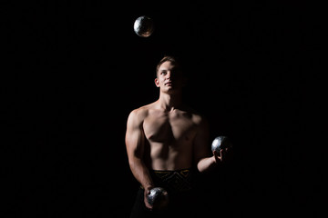 Handsome guy with a muscular body juggles with heavy balls on a black background.Power juggler