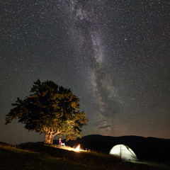 Fototapeta premium Female hiker having a rest at summer night camping in the mountains beside bonfire and glowing tourist tent, sitting on chair under big tree and beautiful night sky full of stars and Milky way.