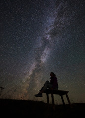 Dark silhouette of female hiker having a rest at summer night in the mountains. Happy woman traveller sitting on a bench, enjoying view of night sky full of stars and Milky way.