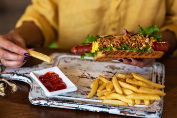 Woman is holding hot dog fully loaded with assorted toppings. Young girl is eating fast food. American meal with french fries on rustic wooden board. Selective focus. Toned image.