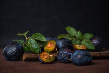 Fresh organic plums with green leaves and in drops. Whole and a half of blue plums fruit on dark vintage background. Toned image. Selective focus.