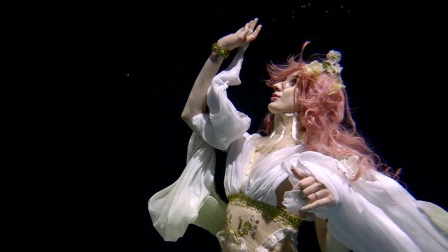 A beautiful girl with pink hair and Eastern attire is underwater on a black background. Looks up and touches the surface of the water.
