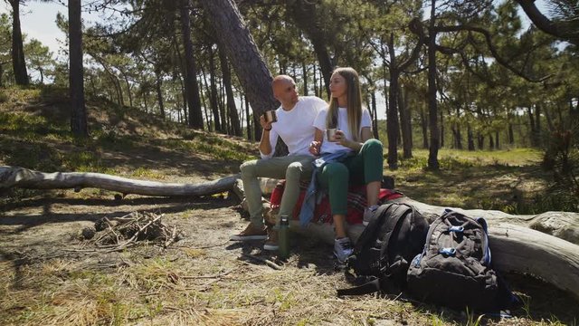 Young couple drinking from metal cups in forest. Happy mana and woman resting on log and drinking hot beverage during hiking outdoor. Recreational pursuit concept