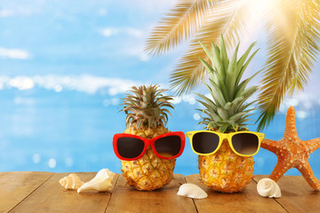 Ripe couple pineapple in stylish sunglasses over wooden table or deck against blue sea, relaxing. Tropical summer vacation concept.