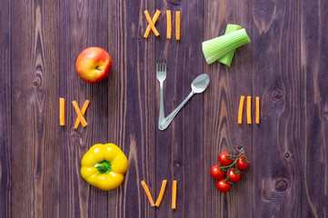 food clock with Roman numerals. Spoon with fork clock hands, vegetables, strawberries, two eggs, mushrooms, Apple, celery on wooden background.
