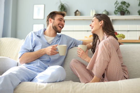 Beautiful young couple in pajamas is looking at each other and smiling on a sofa in the living room.