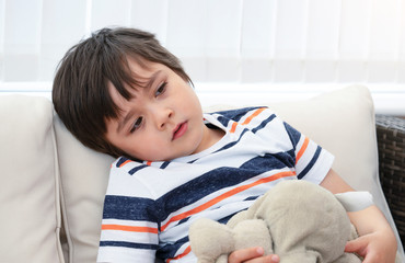 Portrait of little boy sitting in sofa playing teddy bear with bored face, Bored Child with unhappy face sitting with brown bear in conservatory with bright light on summer.