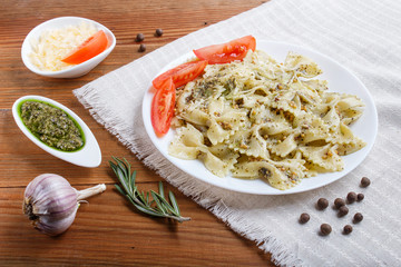 Farfalle pasta with pesto sauce, tomatoes and cheese on a linen tablecloth on brown wooden background.