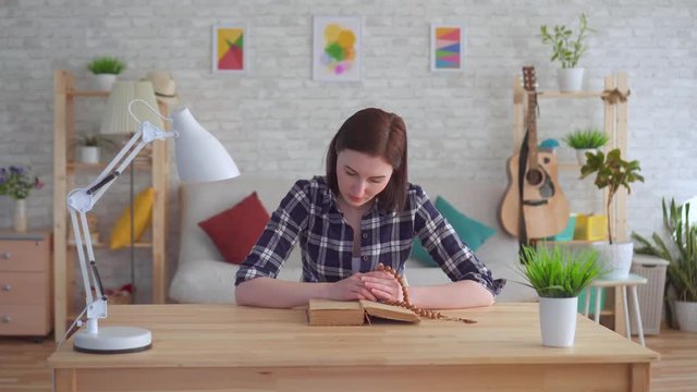 Young woman sitting at a table with a rosary in her hands and reading a book