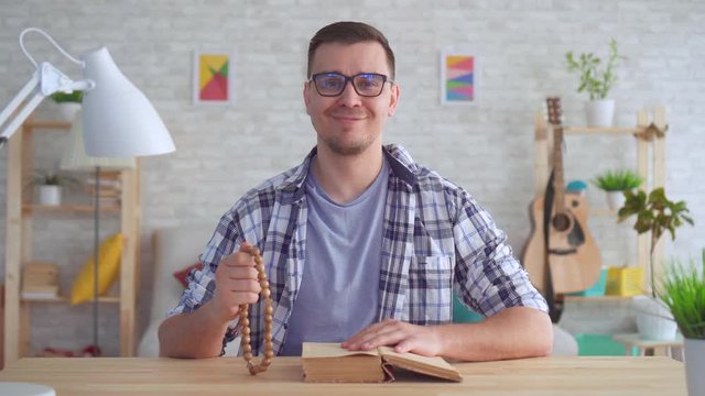 Believing young man with a rosary in his hands and with a Bible on the table looks at the camera