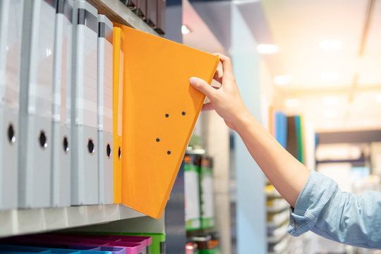 Male hand choosing new orange ring binder file folder from colorful shelf display in stationery shop. Buying office supplies concept