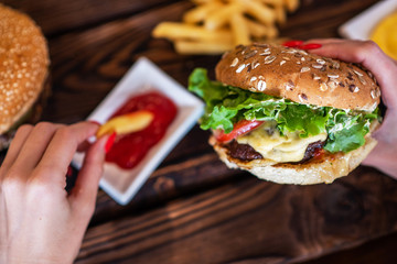 Woman's hand dipping french fries in barbecue sauce. Delicious fresh burger with tomato and cheese. Fast food concept. Beef burger in hands with french fries on rustic wooden board.