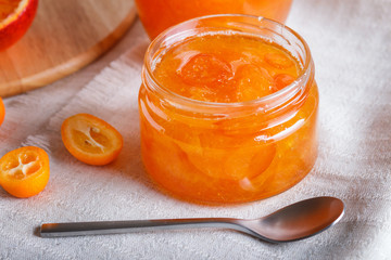 Orange and kumquat jam in a glass jar with fresh fruits on a white linen tablecloth.
