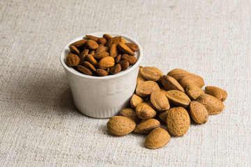 Peeled almonds in a white bowl and almonds nut in shell on beige fabric texture