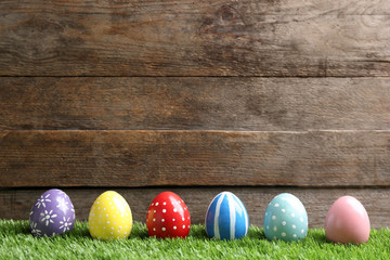 Painted Easter eggs on green grass against wooden background, space for text