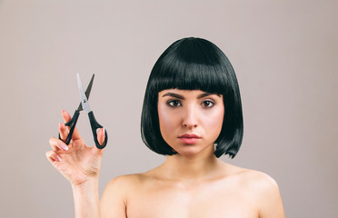 Obraz premium Young woman with black hair posing on camera. Looking straight. Holding scissors in hand. Serious brunette with bob haircut. Isolated on light background.