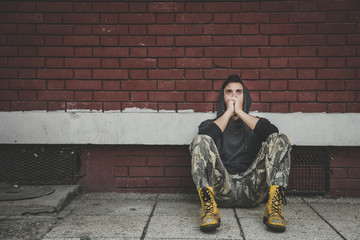 Homeless, Homeless man drug and alcohol addict sitting alone and depressed on the street leaning against a red brick building wall feeling anxious and lonely, social documentary concept 