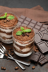 two portions Classic tiramisu dessert in a glass and chocolate bar on dark concrete background