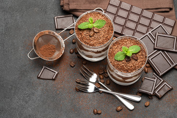 two portions Classic tiramisu dessert in a glass and chocolate bar on dark concrete background