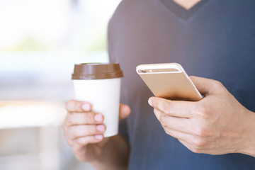 Young male using on cell phone stand in watching message on mobile phone during break hand holding Paper cup of take away drinking coffee hot on the morning in public park.