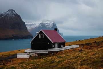 Typical Faroese black wooden house with colourful roof in the village Viðareiði with snow-covered peaks of Kunoy and Kalsoy island (Faroe Islands, Denmark, Europe)