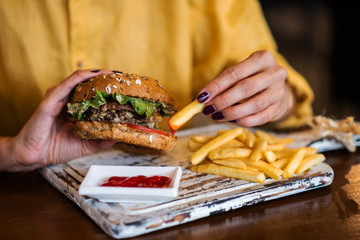 Tasty grilled made burger with beef, tomato, cheese, cucumber and lettuce. Woman holds burger in hands.
