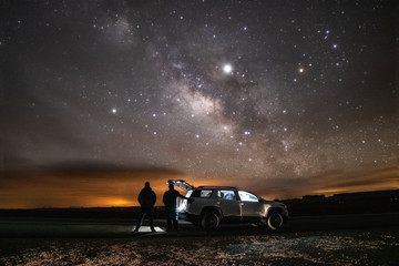 Two silhouettes of people standing under  the Milky Way stretching across the night sky. Adventure scene next to a vehicle in a remote location. 
