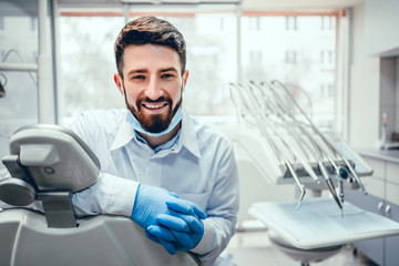 Front view of professional male dentist in white doctor coat and protective gloves sitting in dental chair and equipment, looking at camera and smiling. Bearded man posing during working process.
